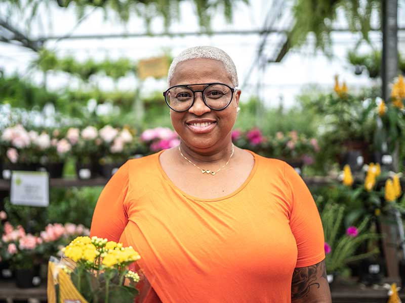 A woman in an orange t-shirt stands in a greenhouse holding yellow flowers