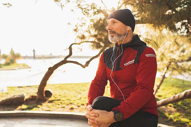 A man clutches his knee in the middle of his outdoor workout. He's running in cold weather, wearing a hat and a red jacket.