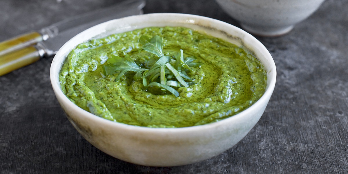 Green chive pesto in a white bowl on granite counter with butter knives and cloth napkins in background.