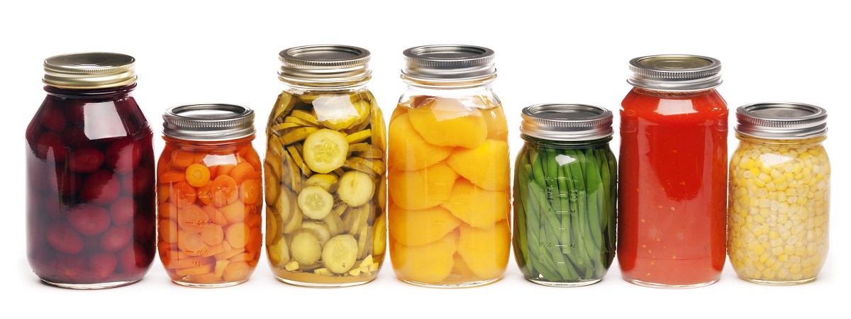 Seven glass jars that contain from left to right, beets, carrots, pickles, peaches, green beans, tomato sauce, and corn.