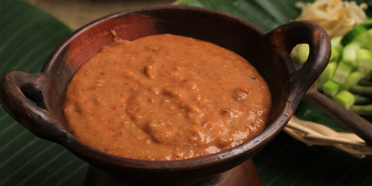 Peanut dressing in small bowl on a large leaf lining a wooden table. Woven baskets in background filled with veggies.
